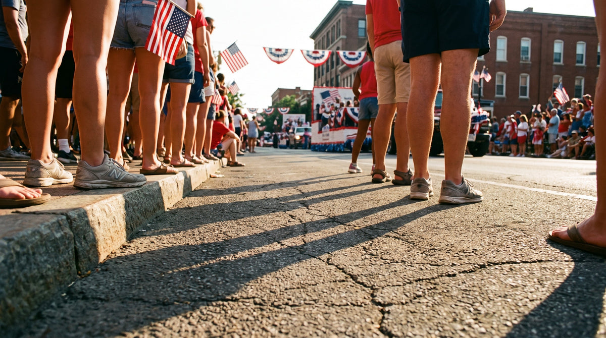 Dense July 4th parade crowd standing on hot city sidewalk — sneakers and sandals on asphalt, American flags in background